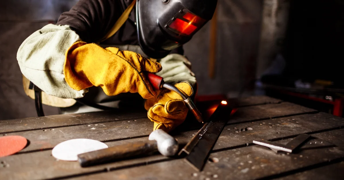A welder wearing protective gear, including gloves and a helmet, uses a welding torch to join metal pieces in a workshop.
